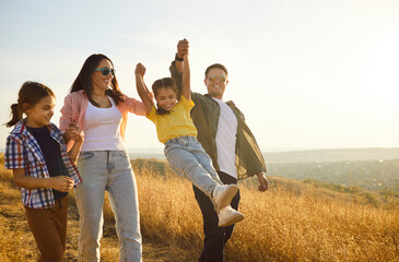 Joyful family walking nature meadow at sunset. Parents swing their laughing child between them as children cheer, enjoying summer light, shared joy together. Concept family unity and love.