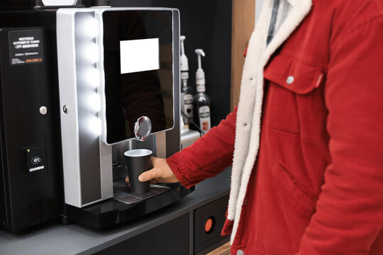 Young man in a red corduroy jacket using a modern, automatic coffee machine to dispense a hot beverage into a black cup.