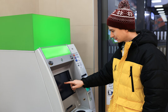 Young man in a bright yellow winter jacket and hat operating an ATM touchscreen, withdrawing or depositing money. - Powered by Adobe