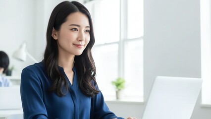 Young Asian woman with long dark hair wearing a blue top smiling and looking at a laptop computer in a bright office environment