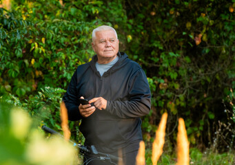 An elderly gray-haired man uses a smartphone while cycling through the forest.