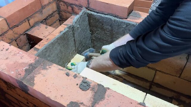 A man is laying the brickwork for a fireclay heating stove core. He is adjusting the masonry with a small hammer