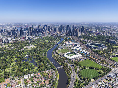 Aerial view of the Yarra River snakes through lush green parks towards the towering city skyline and iconic stadiums, Melbourne, Victoria, Australia.