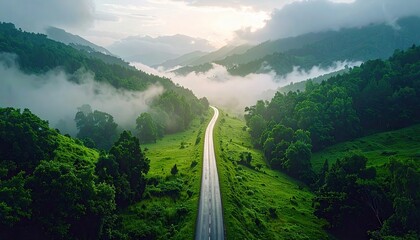Aerial view of a road winding through lush green mountains, with fog in the valleys and sunlight breaking through the clouds.
