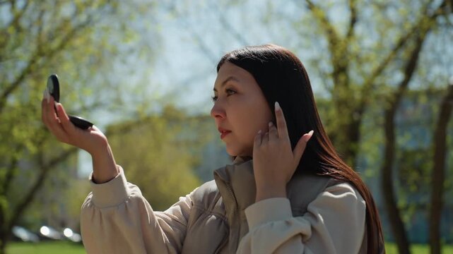 someone enhancing natural beauty beneath sunlight, park scene showing woman doing quick beauty touchup outdoors, individual with compact mirror adjusting makeup amidst sunlight and park surroundings