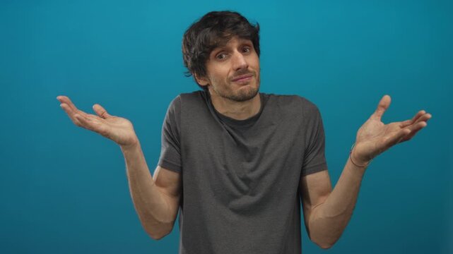 Man wearing grey shirt shrugging open palms with neutral expression showing bare forearms in blue studio; uncertainty.