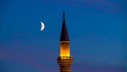 A beautifully illuminated minaret of a mosque stands tall against a deep blue twilight sky with a crescent moon visible.