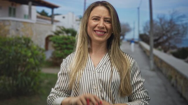 Woman making hashtag gesture with interlocked fingers on coastal street by stone wall and seaside walkway, smiling broadly; playful.