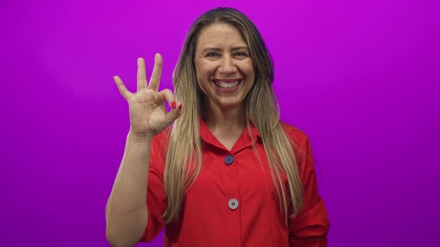 Woman smiling broadly and making ok sign with right hand centered in bright purple studio setting; approval.