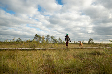 Fototapeta premium Woman hiker in marshy autumn peat together with dog, stepping on wooden trail over raised sphagnum bog. Female explorer pet owner on weekend morning walk at wetland with pet on cloudy fall day.