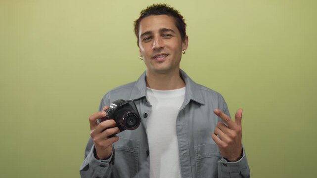 Young hispanic man holding camera in front of isolated yellow background wall with confident pose, showcasing his interest in photography and creative expression.