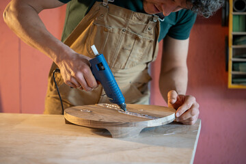 Carpenter working with hot glue gun on cracked surface of wooden stool base, creating border around defect with liquid silicone, next step is filling crack with epoxy resin, workshop creating process.