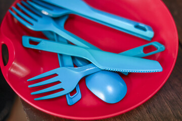 Close up of blue plastic cutlery including fork, spoon, and knife inside red bowl. These reusable utensils are ready for picnic or camping trip, simple and colorful
