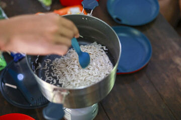 Happy camper cooking instant noodle in pot on portable stove. This simple meal on wooden table represents an outdoor lifestyle, travel, and adventure on summer holiday
