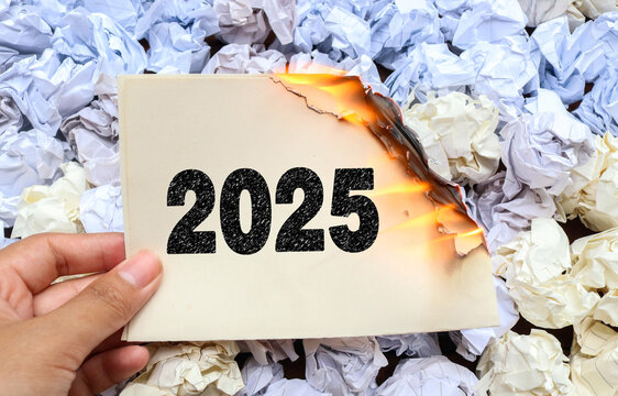 A hand holding a half burned piece of paper with the number 2025 written in a charcoal texture effect, surrounded by crumpled white papers. Goodbye 2025 Concept and new year 2026 concept. End of 2025