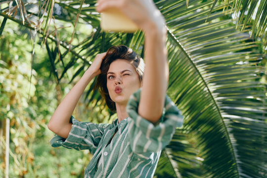 A woman in a striped shirt frames a playful selfie beneath tropical greenery, puckering lips with a kissy expression.