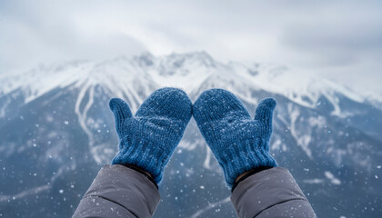 A pair of knitted blue mittens are held up playfully while someone enjoys a peaceful winter landscape