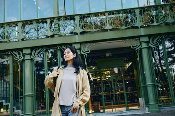 Woman wearing a beige coat on a city street in front of a green storefront illustrates urban...
