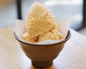 Light peach-colored ice cream scoops in a waffle cone, served in a dark bowl on a light wooden surface