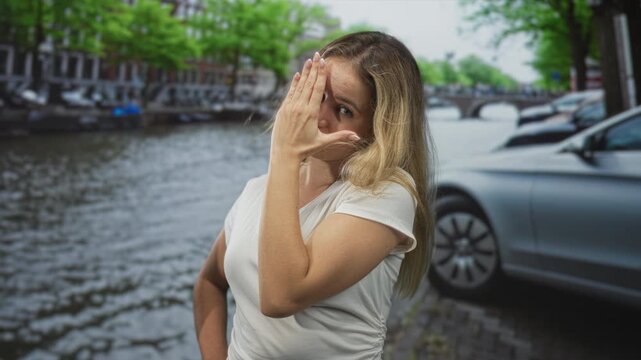Young blonde woman covering eyes with hand while standing beside canal on cobbled street in amsterdam; shyness.