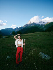 Fototapeta premium Hiking woman stands on a grassy alpine meadow with a trekking staff, red pants and white top, mountains in the background and a clear sky for a serene outdoor adventure