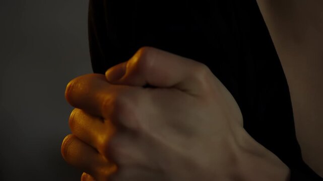 Intimate cinematic close-up of a shoulder and clasped hand in soft, low key lighting, revealing skin texture, collarbone contours, and warm amber highlights against dark fabric.