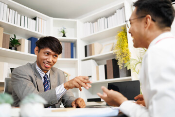 Obraz premium Two men are sitting at a desk, one of them is holding a piece of paper. They are having a conversation, and it seems like they are discussing something important. The atmosphere is tense 