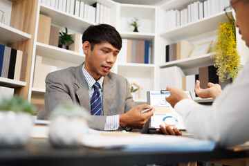 Two men are sitting at a desk, one of them is holding a piece of paper. They are having a conversation, and it seems like they are discussing something important. The atmosphere is tense
