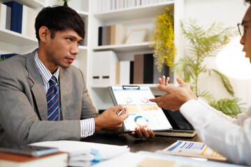 Two men are sitting at a desk, one of them is holding a piece of paper. They are having a conversation, and it seems like they are discussing something important. The atmosphere is tense
