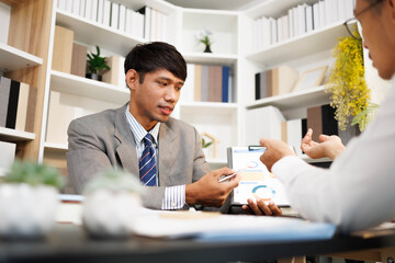 Two men are sitting at a desk, one of them is holding a piece of paper. They are having a conversation, and it seems like they are discussing something important. The atmosphere is tense
