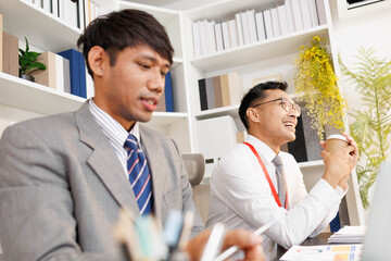 Two men are sitting at a desk, one of them is holding a piece of paper. They are having a conversation, and it seems like they are discussing something important. The atmosphere is tense
