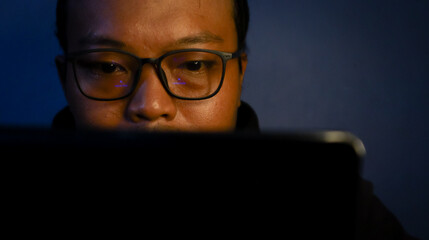 Focused Asian man working late in front of laptop with warm and moody lighting, creative freelance concept.