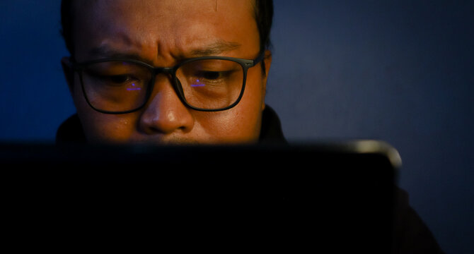 Focused Asian man working late in front of laptop with warm and moody lighting, creative freelance concept.