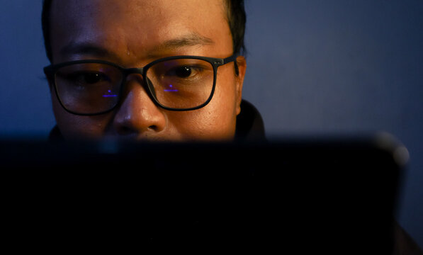 Focused Asian man working late in front of laptop with warm and moody lighting, creative freelance concept.