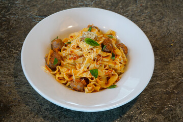 Fresh tagliatelle pasta with Italian sausage, tomato sauce, basil, and grated cheese served in a white bowl on a rustic stone background, captured in natural light for authentic food styling.