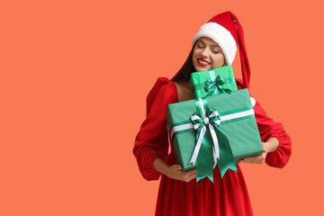 Happy young woman in dress and Santa hat with gift boxes on orange background. Christmas concept