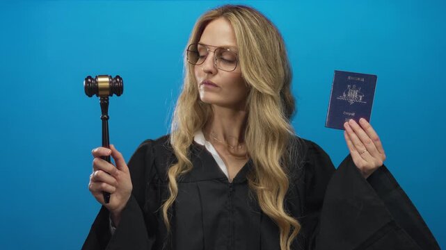 Woman judge in black robe holds an australian passport and gavel against a blue isolated background, symbolizing law and immigration.