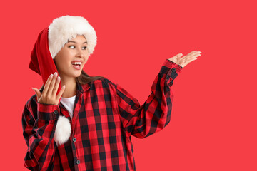 Beautiful young woman in checkered pajamas and Santa hat on red background. Christmas celebration
