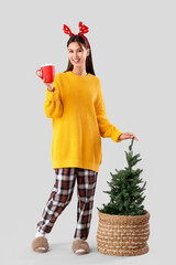Beautiful young woman in pajamas with cup of hot cocoa and Christmas tree on grey background
