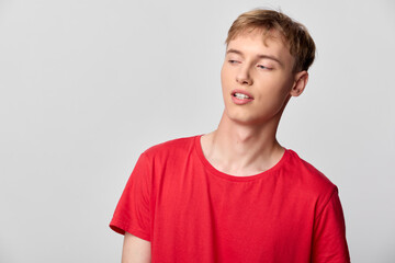 Obraz premium Young man in a red T shirt stands against a neutral backdrop with a relaxed pose and tilted head, conveying casual style, everyday charm, and a candid fashion moment