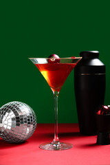 Disco ball, glass of tasty red martini, Christmas balls and bartender tools on red table near green wall