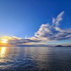The image depicts a serene seascape during sunset, with the sun partially below the horizon casting a warm golden reflection on the calm water. The sky is mostly clear with a few scattered clouds