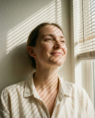 Woman gazing out window with subtle smile, shadows on her face.