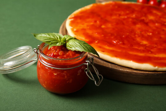 Board with raw dough for pizza and jar of tomato sauce on green background, closeup - Powered by Adobe