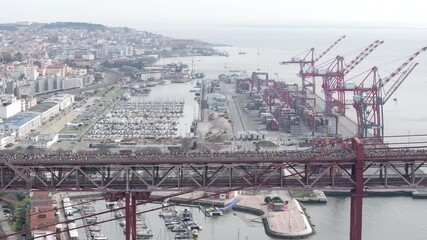 Side-on aerial drone shot of half marathon and 10k runners crossing the iconic red 25th April suspension bridge above the Lisbon waterfront, marina, port docks, and industrial cranes in Portugal