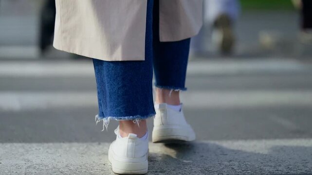 Low-angle close-up sequence showing a pedestrian in white sneakers and frayed-hem denim jeans walking across a striped crosswalk on an urban street, capturing motion and texture.