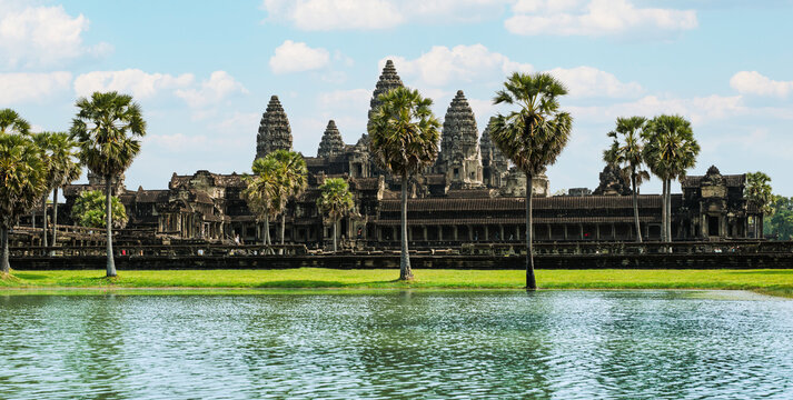 Wide panoramic view of Angkor Wat temple reflection in water moat with palm trees under blue sky with white clouds - Powered by Adobe