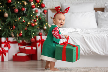 Cute little baby in elf costume and reindeer horns with gift box near Christmas tree in bedroom