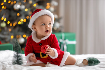 Cute little baby in Santa costume with Christmas ball and mini fir trees sitting on bed in bedroom
