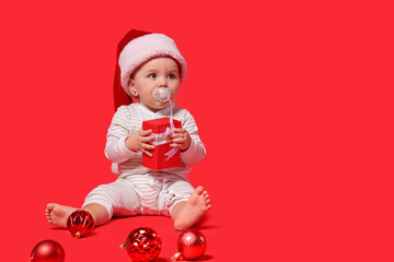 Cute little baby in Santa hat with gift box and Christmas balls sitting on red background